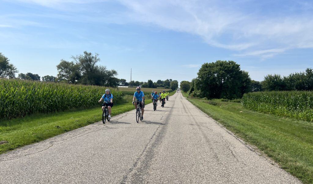 A group of bicyclists approaches on a country road through farm country with some trees in the background under a blue sky with some hazy white clouds.