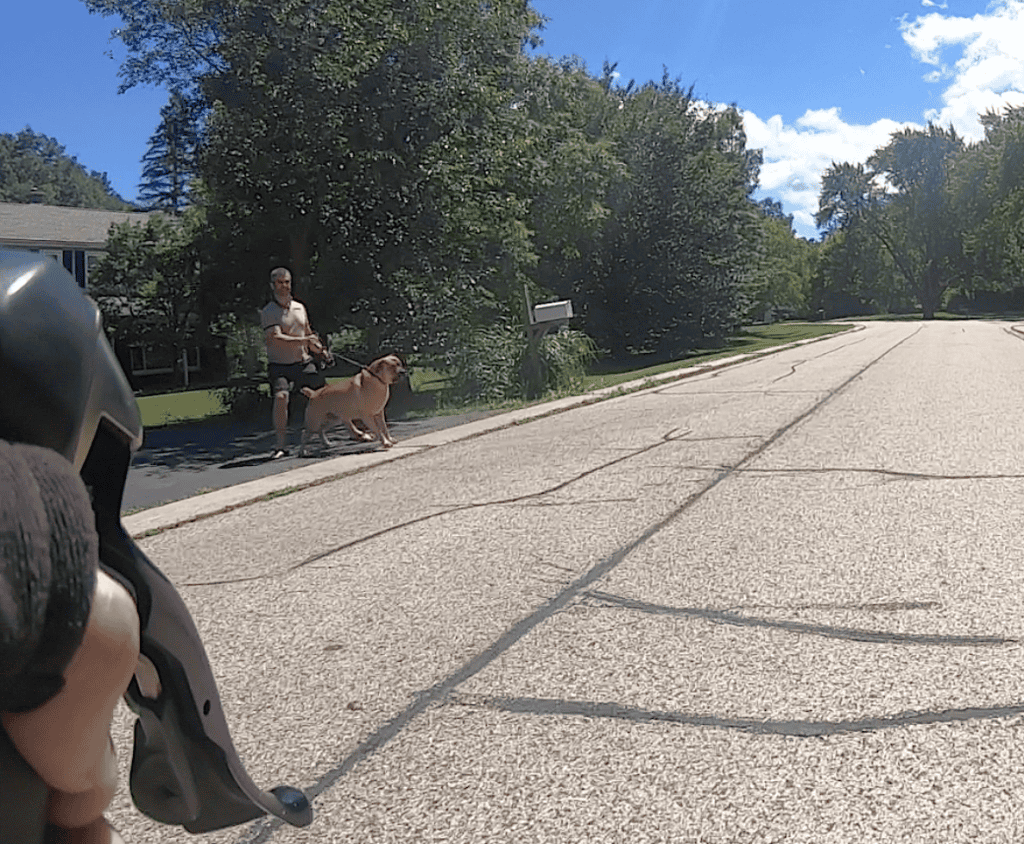 From the handlebar view on a bicycle, a dog owner bracing to hang on to a big dog on a leash appears on the left side of a wide street in a suburban neighborhood on a sunny day.