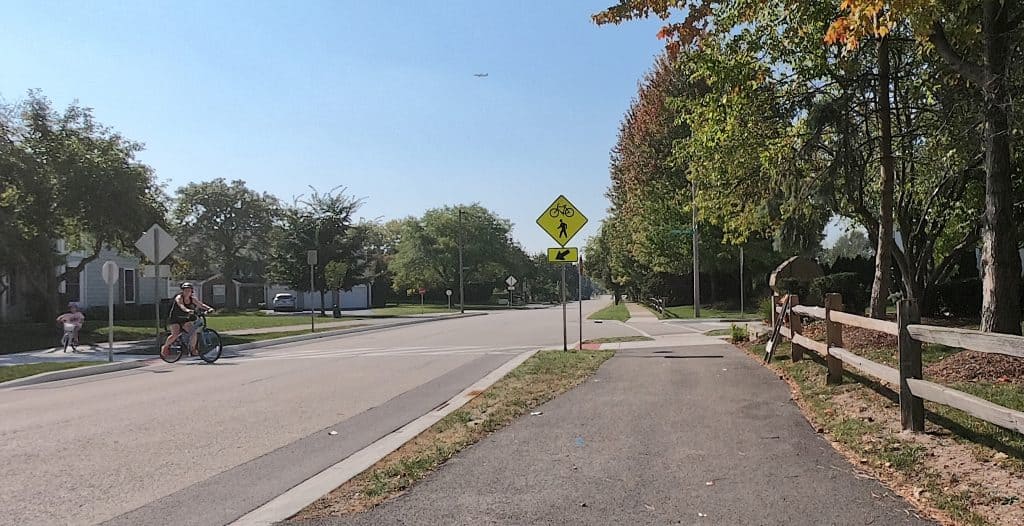 Scenic suburban bike path in Chicago suburbs with bike and pedestrian crossing signs, lush trees, and safe sidewalk for cycling and walking.