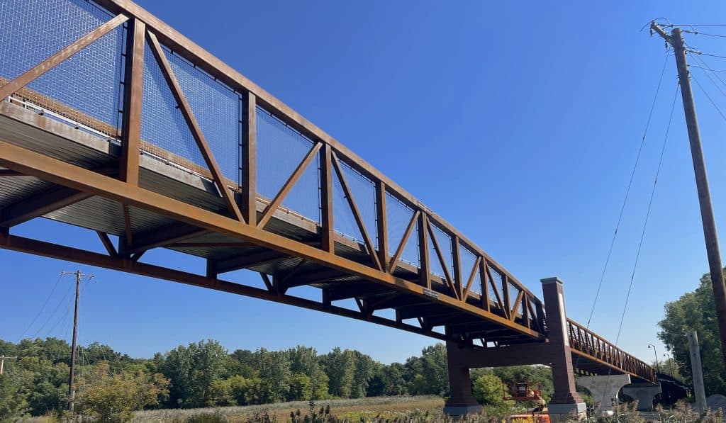 A bridge extends across a road into tree- and prairie-covered land under a blue sky.