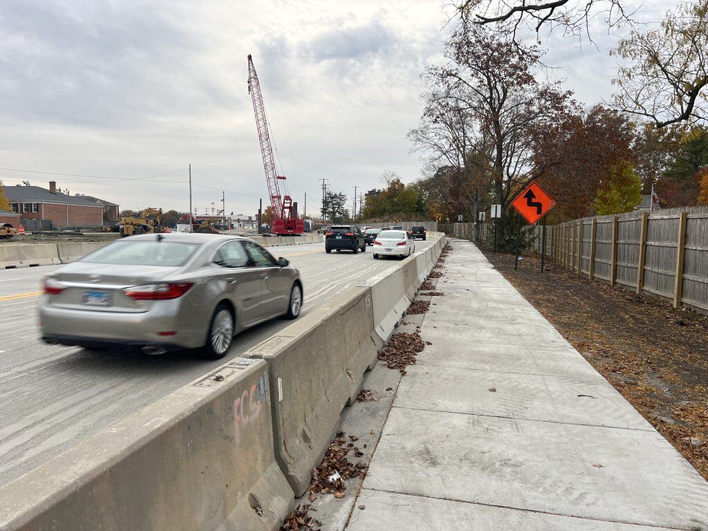 Light gray sidewalk with autumn leaves alongside a busy road, safety barriers, and a construction site in Suburban Chicago.