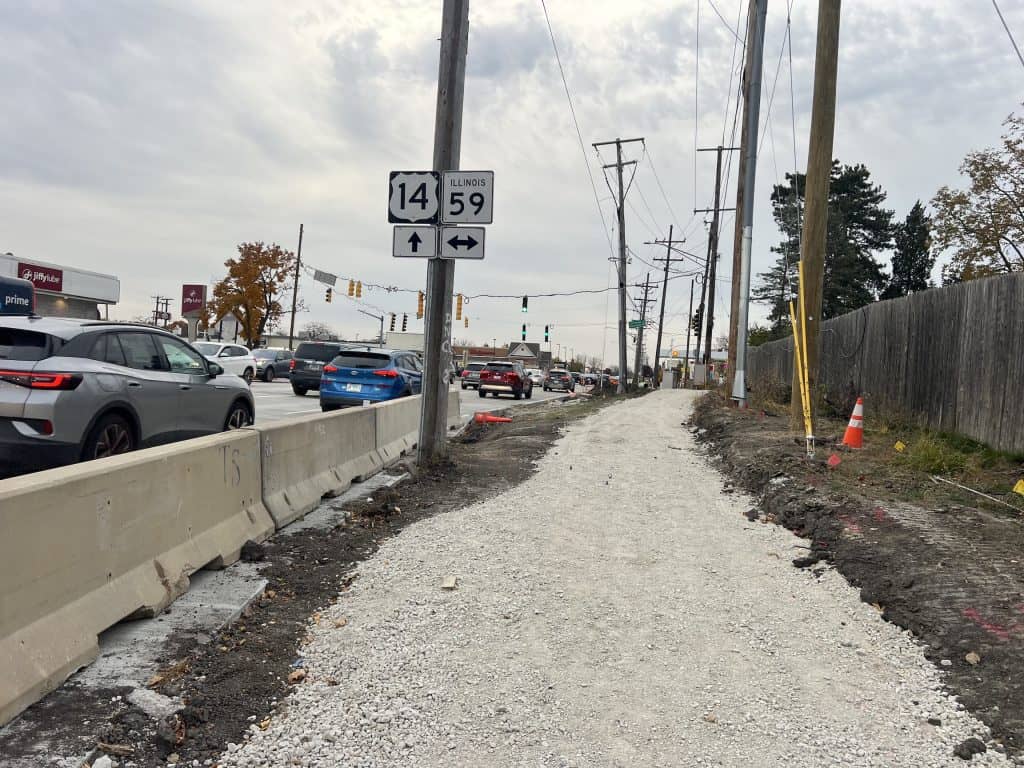 Unfinished bike path along busy road in suburban Chicago with traffic and construction signs.