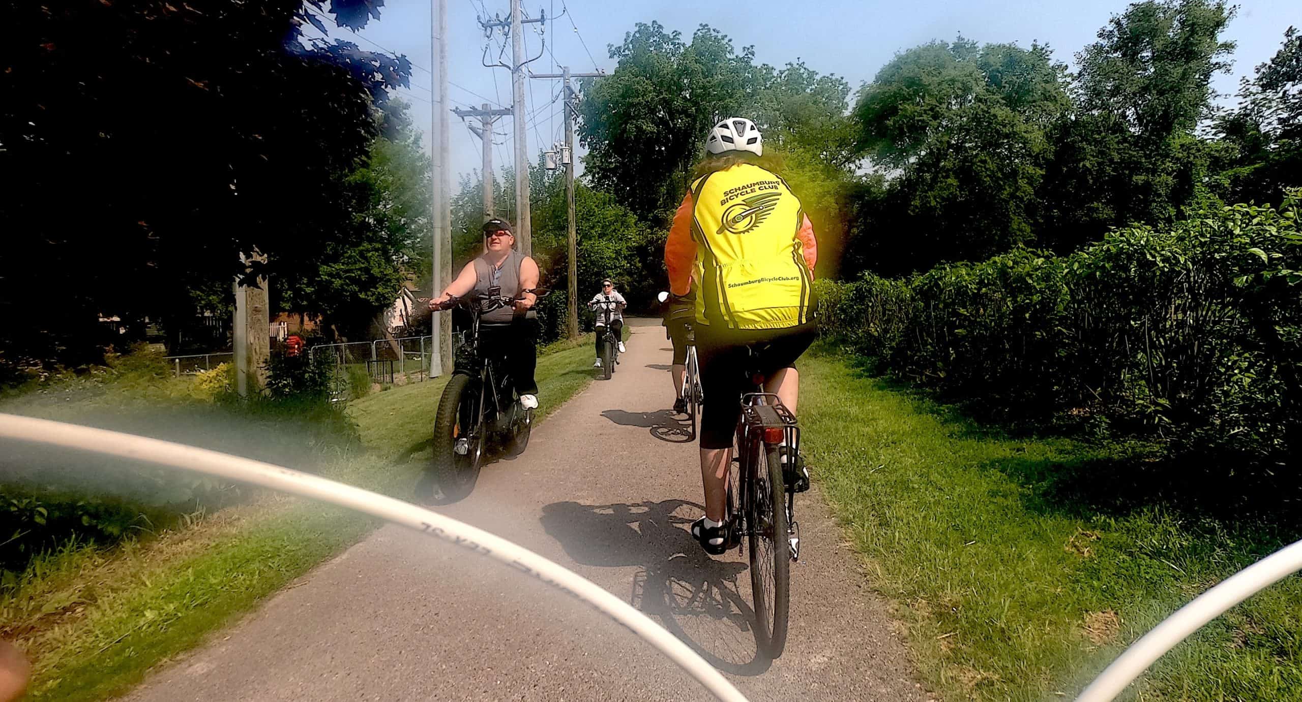Two unhelmeted bicyclists on e-bikes approach our view behind traditional bicyclists on a bush- and tree-lined bike trail.