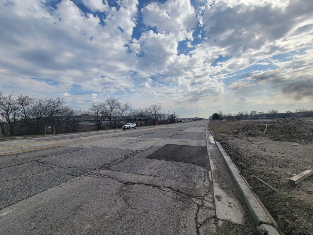 Looking down a wide, beat-up road in an open suburban area under a partly cloudy sky in scrubby winter weather.