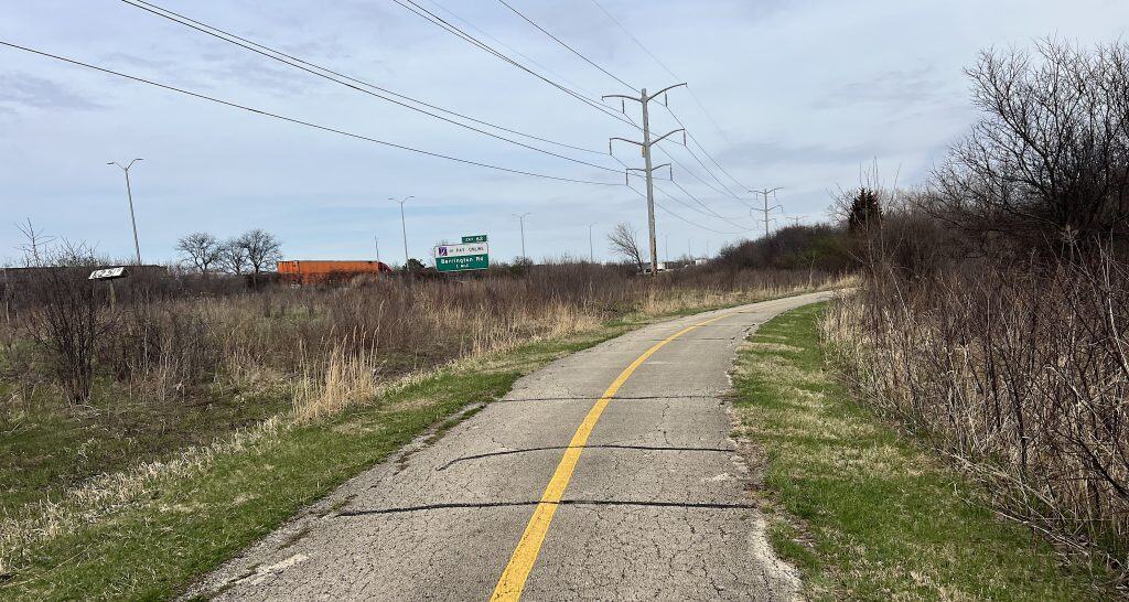 A scenic suburban bike path with overgrown vegetation and utility poles in the background.