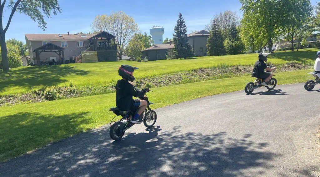 A young child riding a electric bicycle on a suburban street with green lawns and trees.