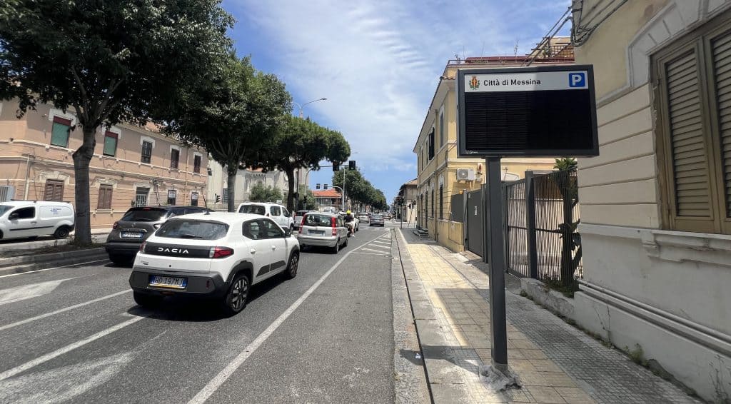 City street with parked cars, trees, and a digital parking sign in Messina, Italy.