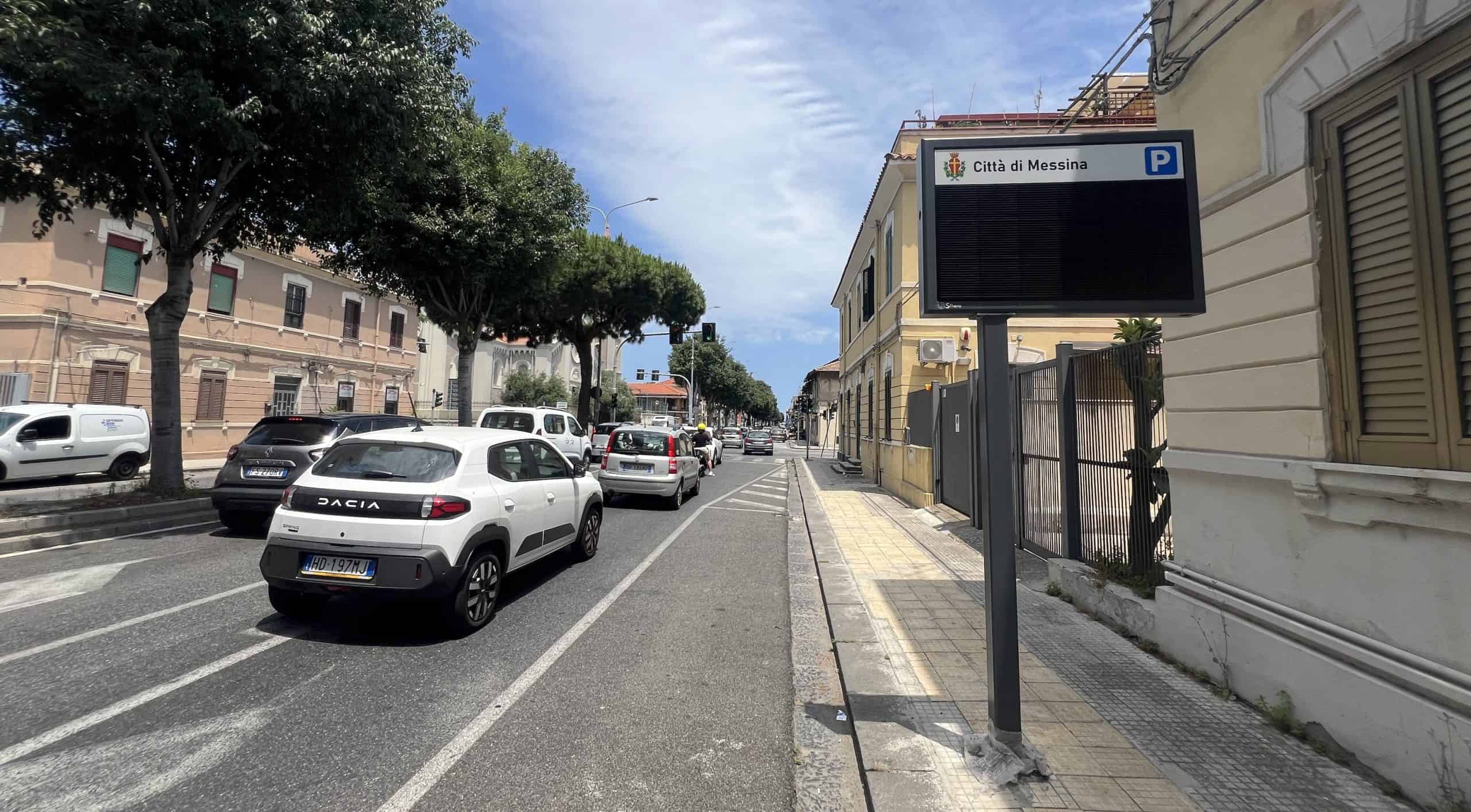 City street with parked cars, trees, and a digital parking sign in Messina, Italy.