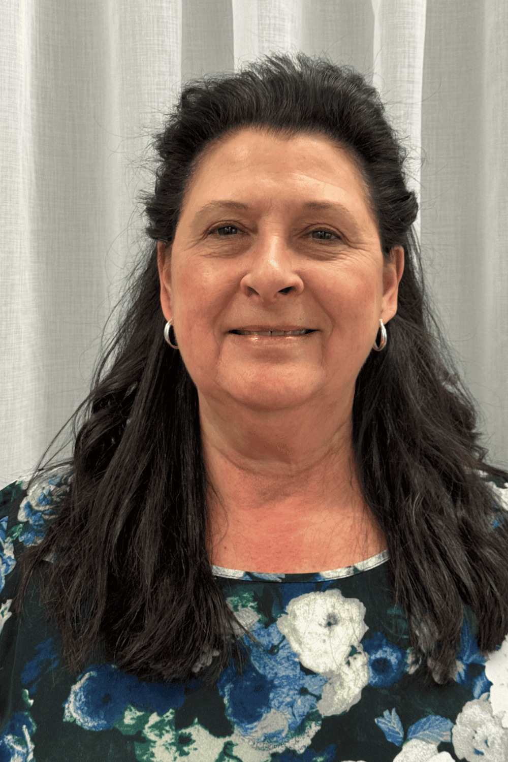 Bright woman with dark hair and floral top smiling in front of window shades.