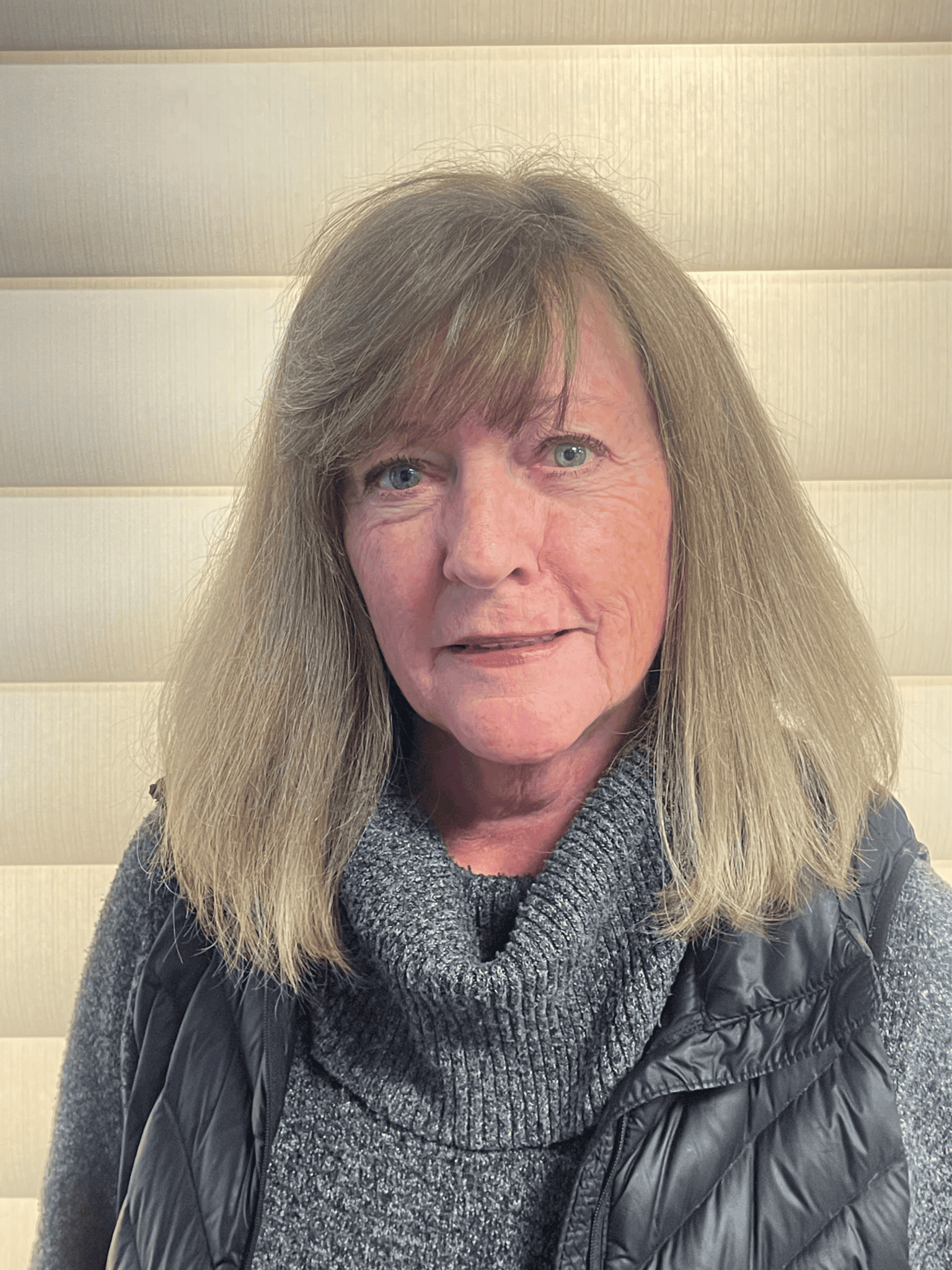 Close-up of a woman with long blonde hair smiling, wearing a grey turtleneck and black quilted vest, indoors.
