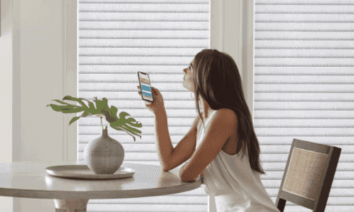 Young woman with long brown hair sitting at a table, holding a smartphone, with white blinds and a potted plant in the background.