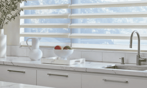 Bright modern kitchen with white cabinets, marble countertop, and a large window showing a blue sky with clouds.