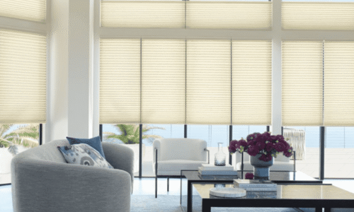 Bright living room with large windows, beige blinds, gray sofa, white armchair, coffee table, and ocean view in the background.
