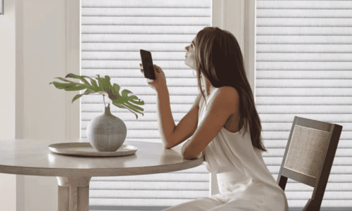 Young woman with long hair in a white dress sitting at a dining table, holding a smartphone, with a decorative vase and plant, bright room with blinds.
