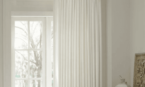 Bright room with white curtains hanging from a dark curtain rod, sunlight streaming through a large window, and a cozy interior setting.