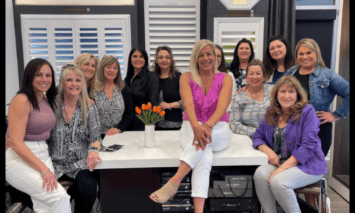 Group of women gathered in a window treatment showroom with shutters and blinds display in the background.