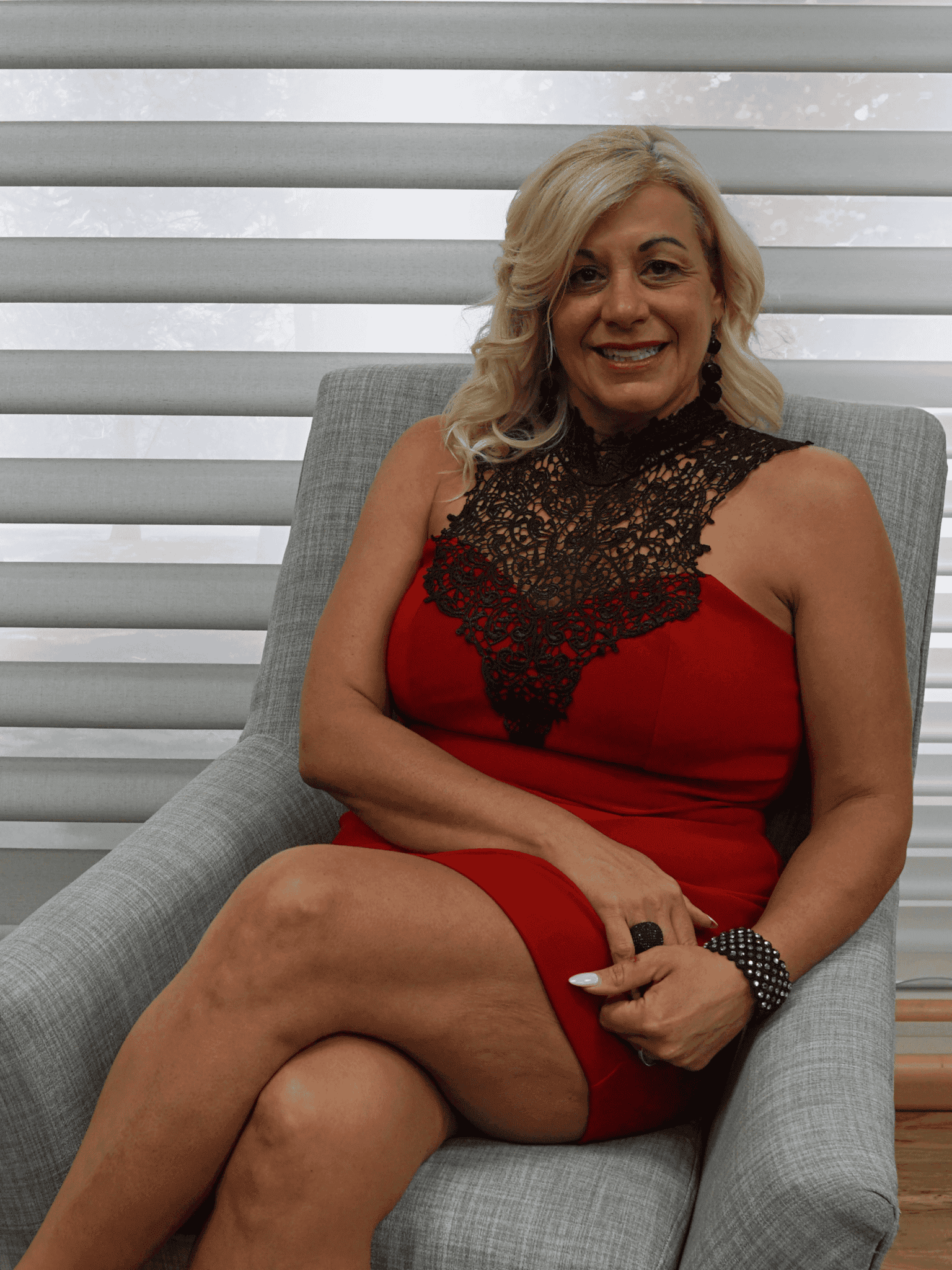 Angie Romano sitting on a gray armchair, wearing a red dress with black lace details, smiling at the camera.