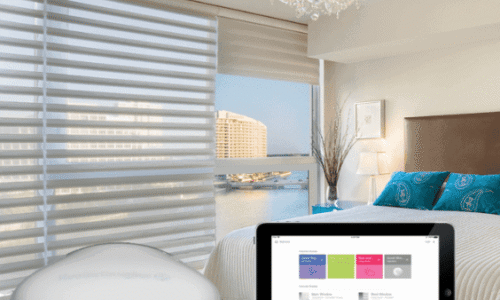 Contemporary bedroom featuring a smart home device on a table, large window with blinds, chandelier, and a tablet displaying a home automation app.