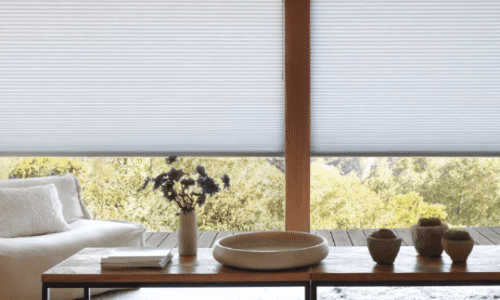 Close-up of white window blinds in a cozy living room with a view of green trees outside.