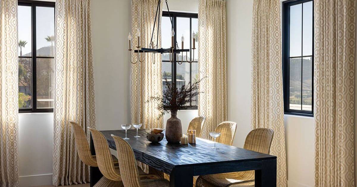 Bright dining area featuring large windows, cream patterned curtains, a rustic black wood table, wicker chairs, and minimalistic decor for a cozy, contemporary look.