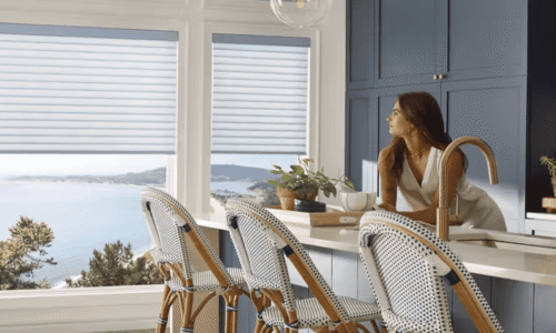 Bright modern kitchen with large windows overlooking ocean, featuring blue cabinets, white countertops, and wicker bar stools.