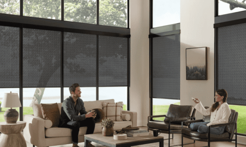Two people sitting in a modern office lounge with large windows, blinds, and contemporary decor, engaging in conversation.
