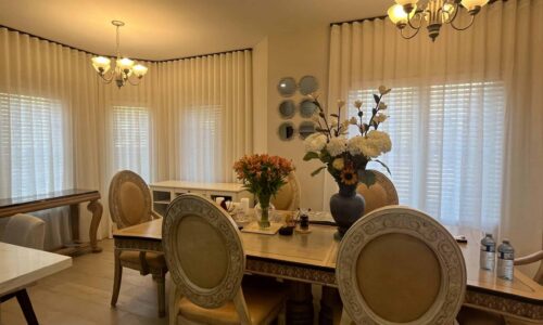 Spacious dining room featuring a large wooden table, six upholstered chairs, floral centerpieces, and elegant lighting fixtures.