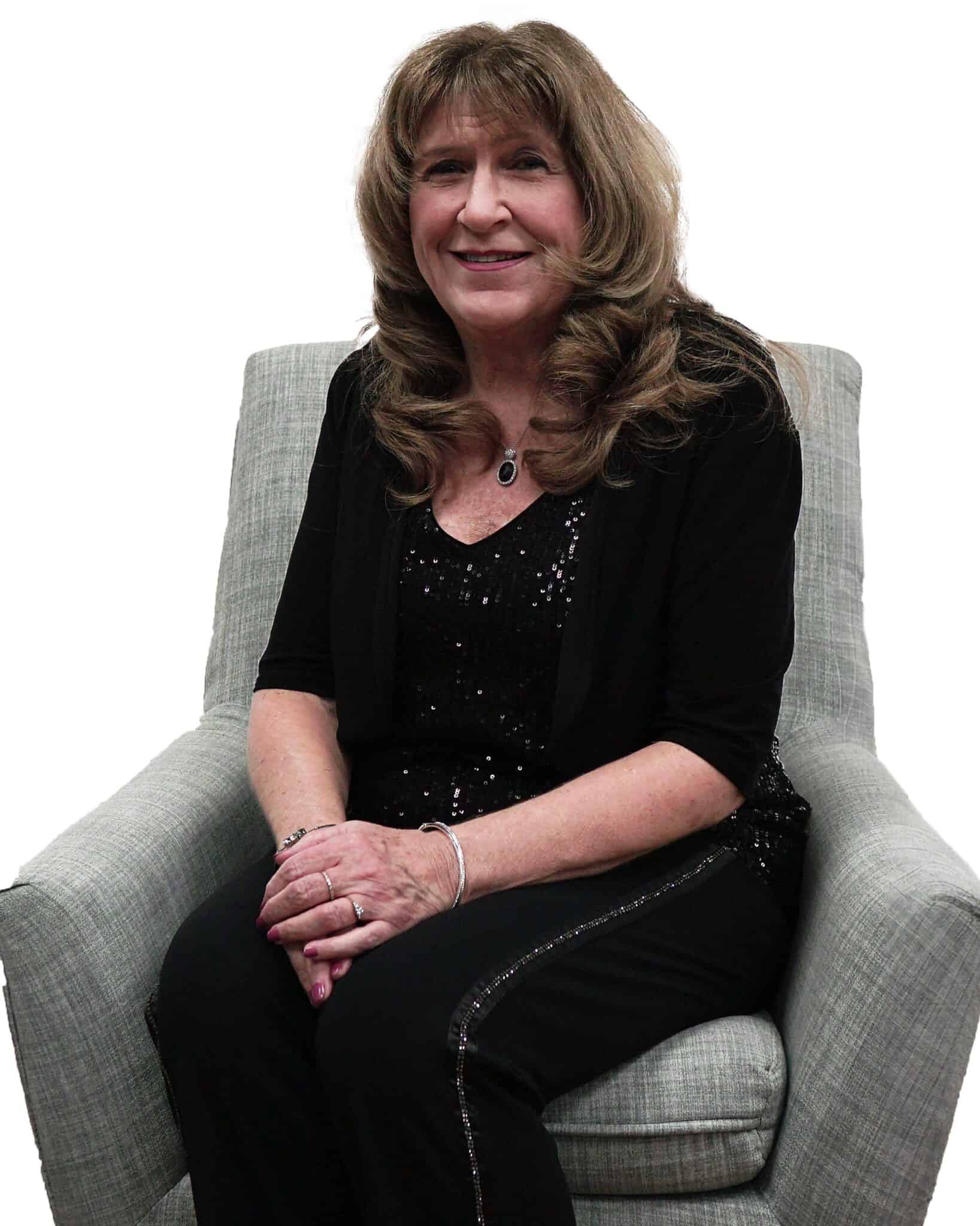 A woman with long, wavy brown hair smiling while sitting in a light gray armchair, wearing a black outfit with a sparkly top and a necklace.