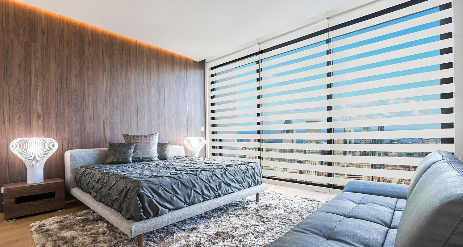 Bright modern bedroom with large windows, wooden wall, gray bed, and cozy seating area, overlooking city skyline.