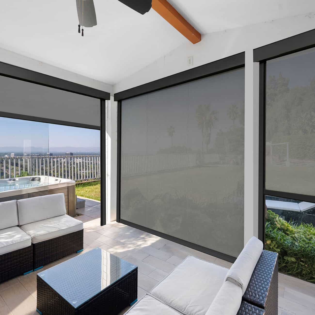 Bright sunroom featuring expansive glass windows with roller shades, overlooking a lush garden and scenic landscape. Comfortable seating area with wicker furniture and a small coffee table.