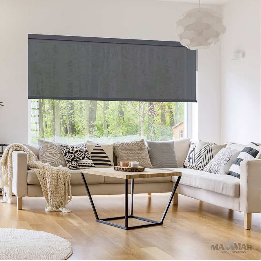 Bright living room featuring a large window with a dark roller shade, beige sectional sofa with patterned cushions, and a minimalist black coffee table. Natural light highlights the airy, inviting spa.