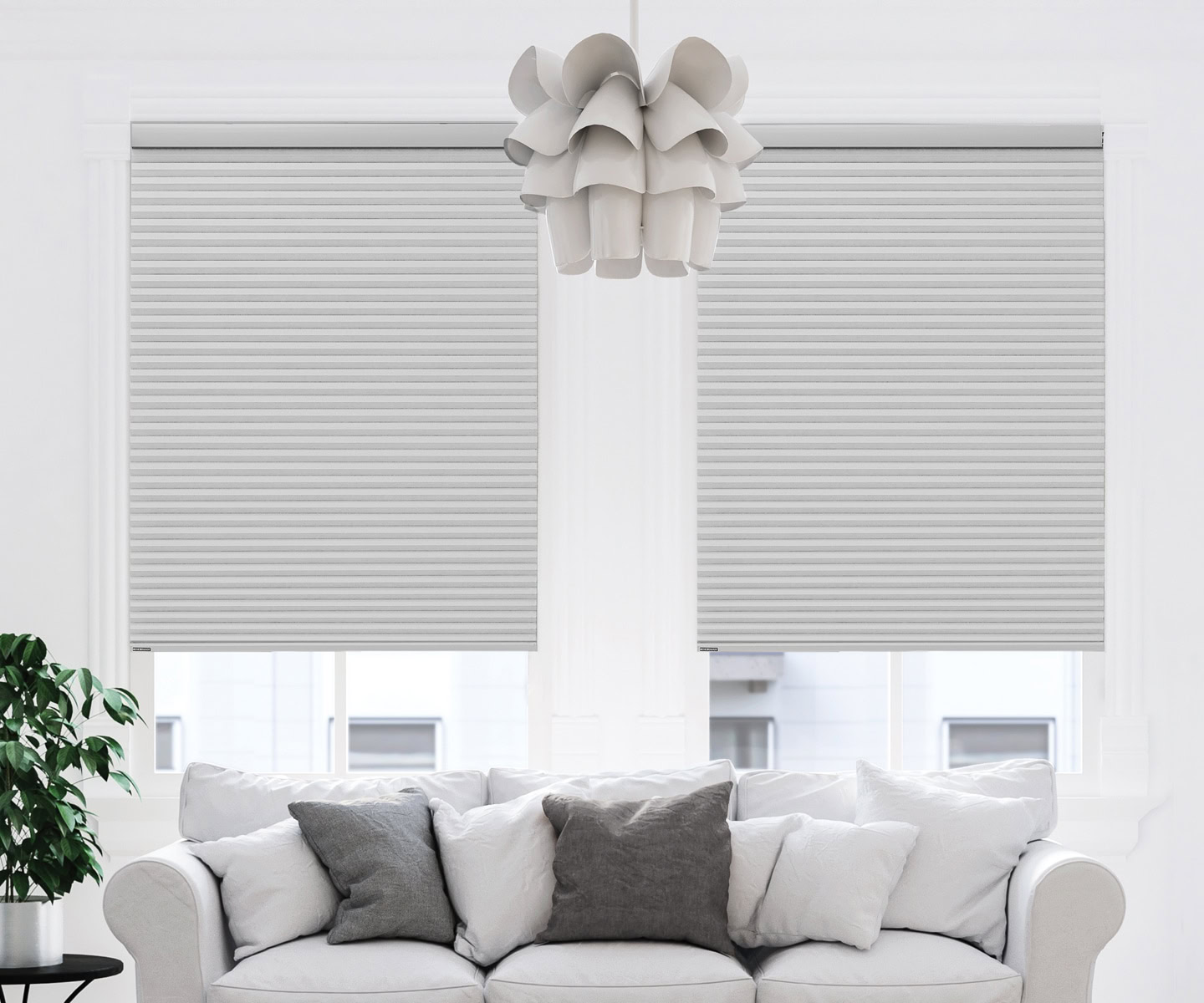 Two white Portrait™ Honeycomb Shades covering large windows in a modern living room with a white sofa and decorative pillows.