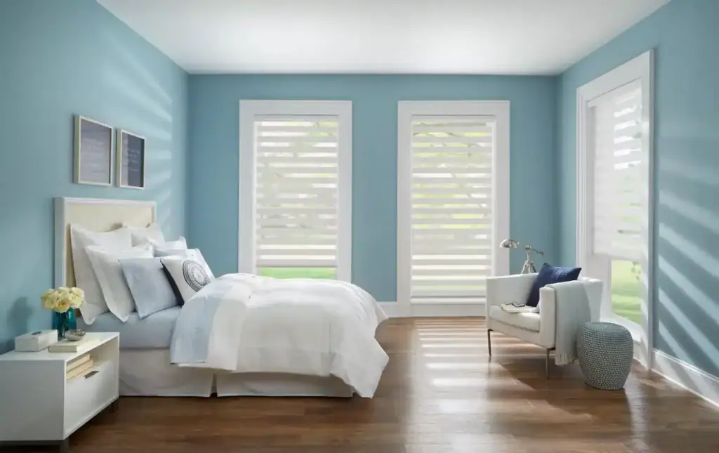 Spacious bedroom with light blue walls, large windows with white blinds, white bed, and cozy seating area with armchair and side table.