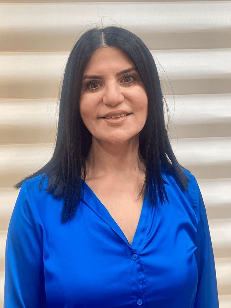 Portrait of a woman with dark hair wearing a bright blue blouse, smiling softly, standing in front of a neutral background.