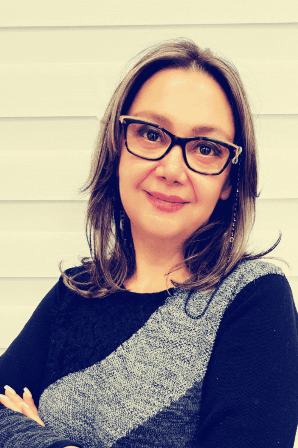Close-up portrait of a woman with glasses, shoulder-length brown hair, smiling, wearing a black and gray sweater, standing against a white wooden background.