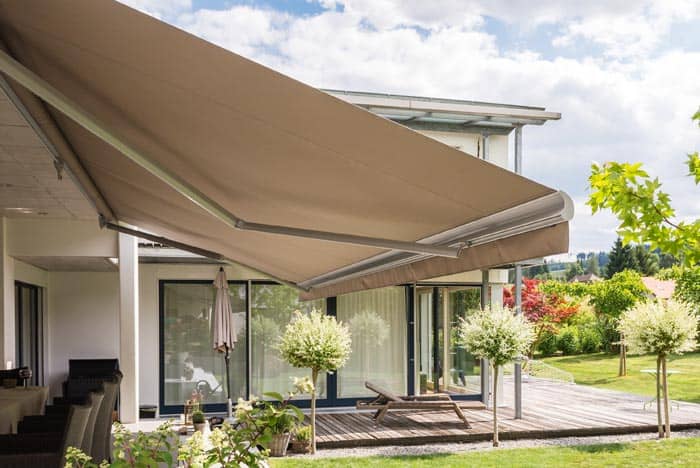 Modern beige retractable awning providing shade over outdoor patio area with lush greenery and trees in the background.