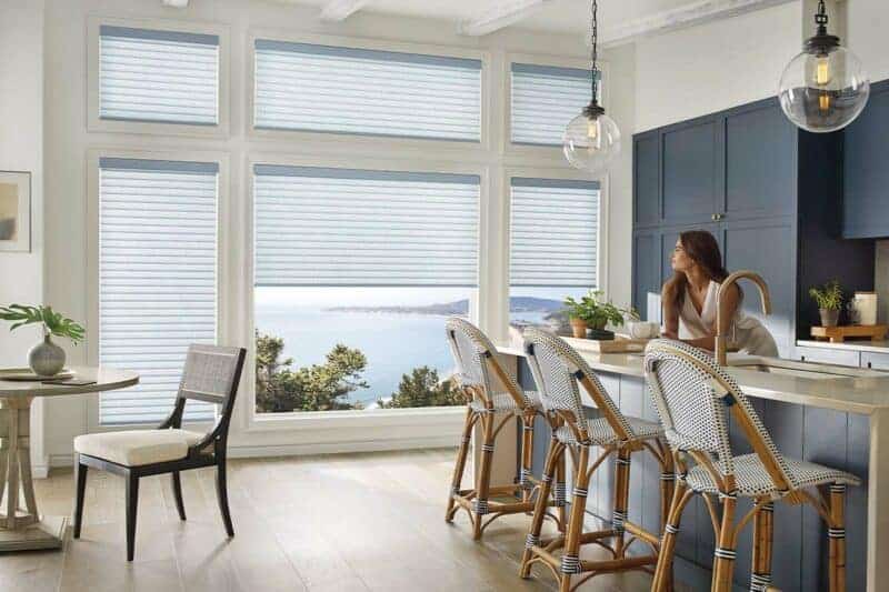 Bright kitchen with large windows covered by white blinds, featuring a woman sitting at the counter, bar stools, and a view of trees and water outside.
