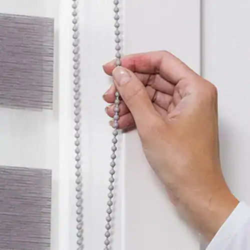 Beaded chain for window blinds control, close-up of hand operating the chain on a modern white shade.