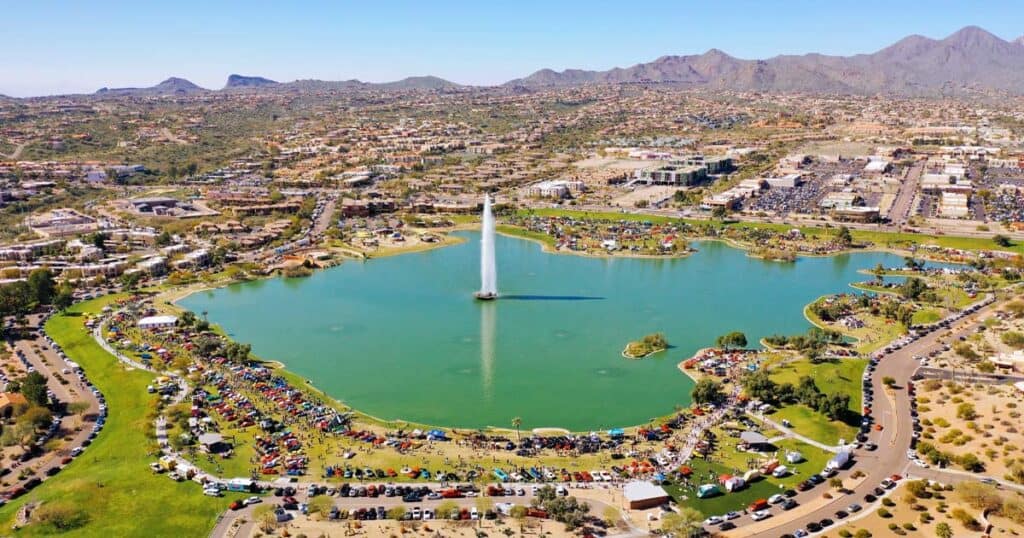 Aerial view of a large city park with a central lake and a fountain, surrounded by parking lots and commercial buildings, with mountain ranges in the background.