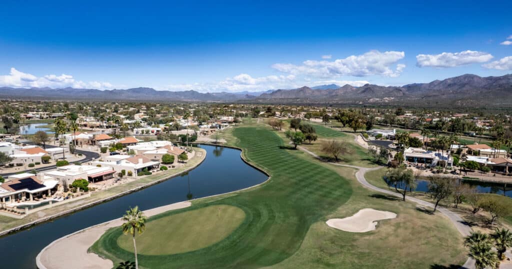Bright, sunny golf course with lush green fairways, sand traps, and water features set against a backdrop of mountains under a blue sky. Perfect for outdoor recreation and leisure activities.