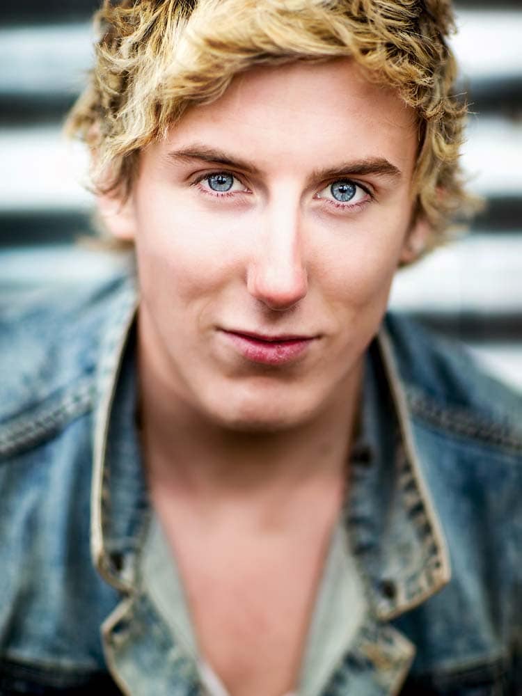 Bright blue-eyed young man with blonde curly hair in a denim jacket, outdoor headshot for professional Sussex headshots and corporate branding.