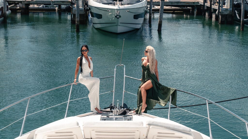 Luxurious women on yacht in marina, high-end lifestyle, fashion, leisure, nautical yacht vacation, premium boating experience, elegant summer attire, scenic harbor backdrop.