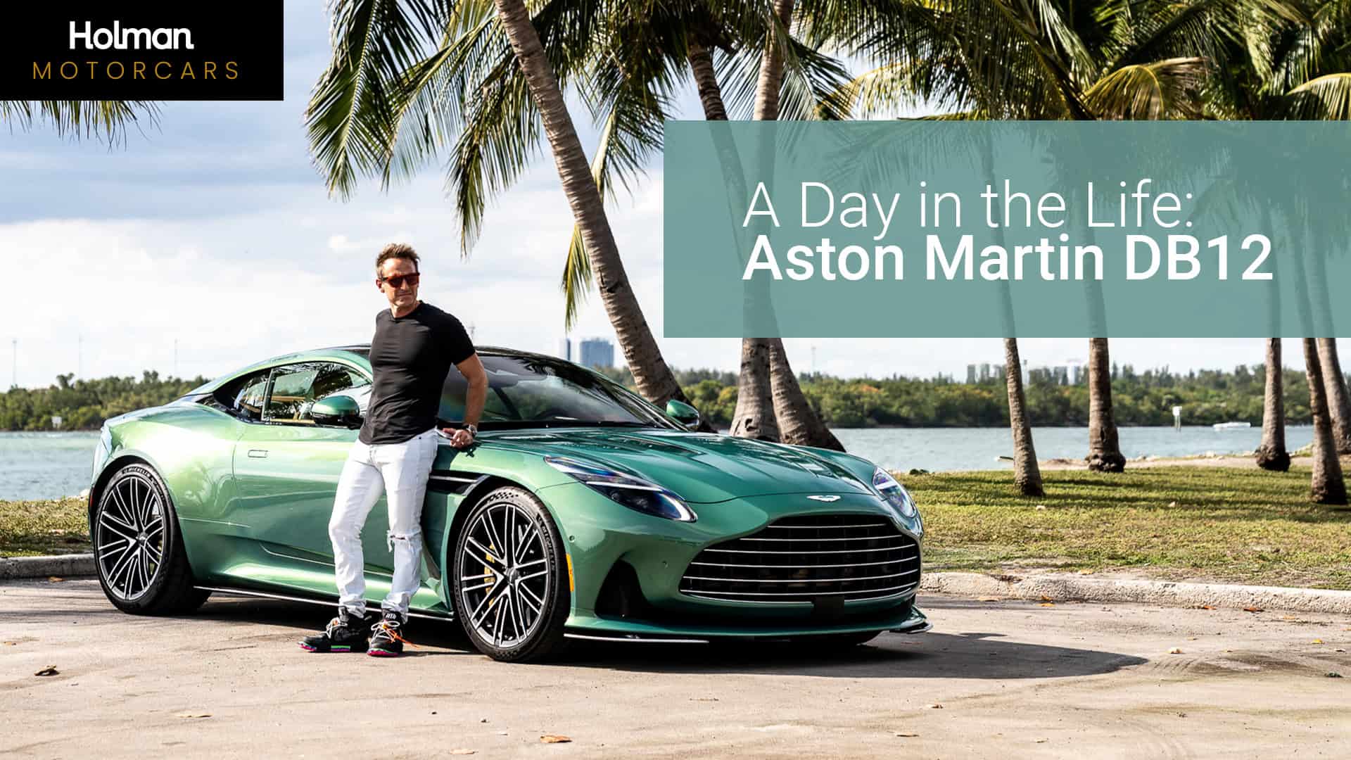 Aston Martin DB12 luxury sports car parked by the water with palm trees, featuring a man in casual attire leaning on the vehicle in a scenic outdoor setting.