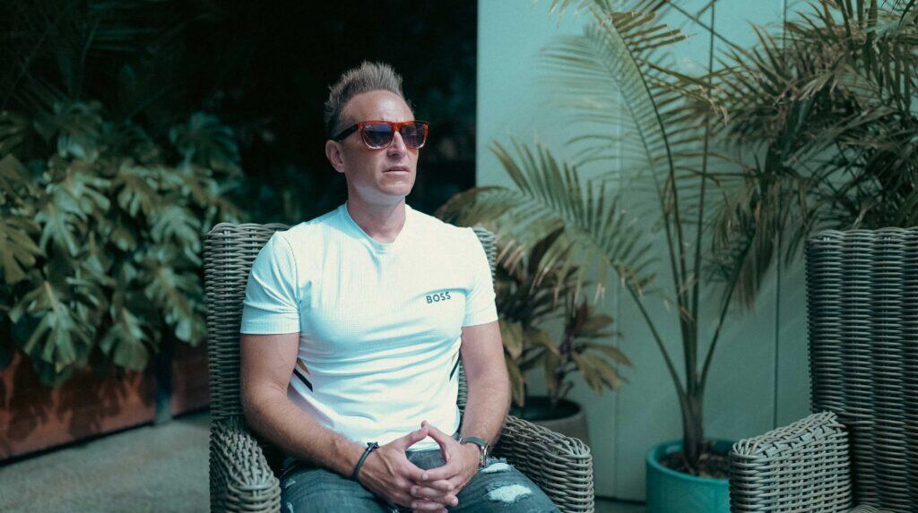 Relaxed man with sunglasses sitting on wicker chair amidst indoor plants, showcasing luxury lifestyle and contemporary outdoor furniture design.