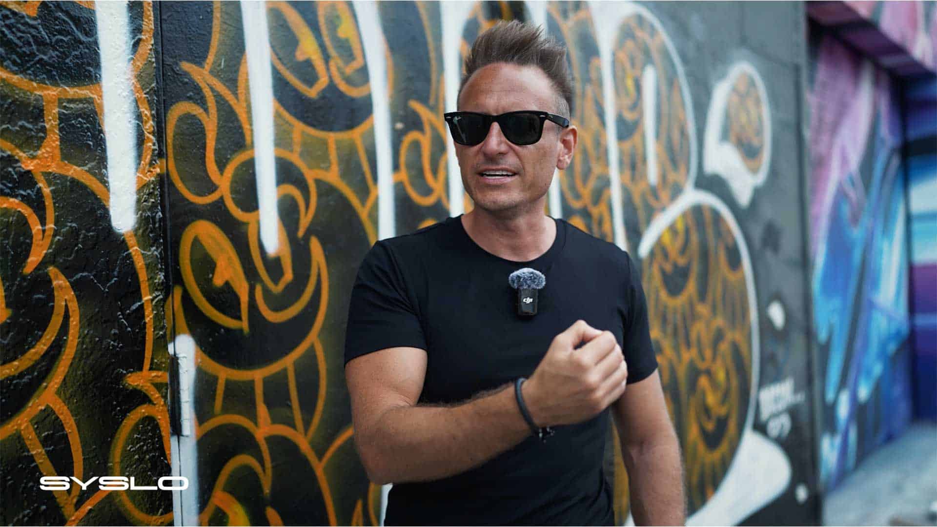 Vibrant urban graffiti art on a wall in a city alley, featuring bold colors and intricate designs, with a man in sunglasses and black shirt speaking confidently, exemplifying creative expression and modern street art culture.