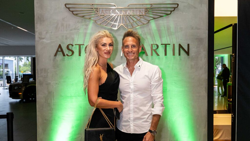 Aston Martin event with a stylish woman and a man posing together in front of the Aston Martin logo at Syslo Ventures. Luxury lifestyle, automotive, and networking in a premium setting.