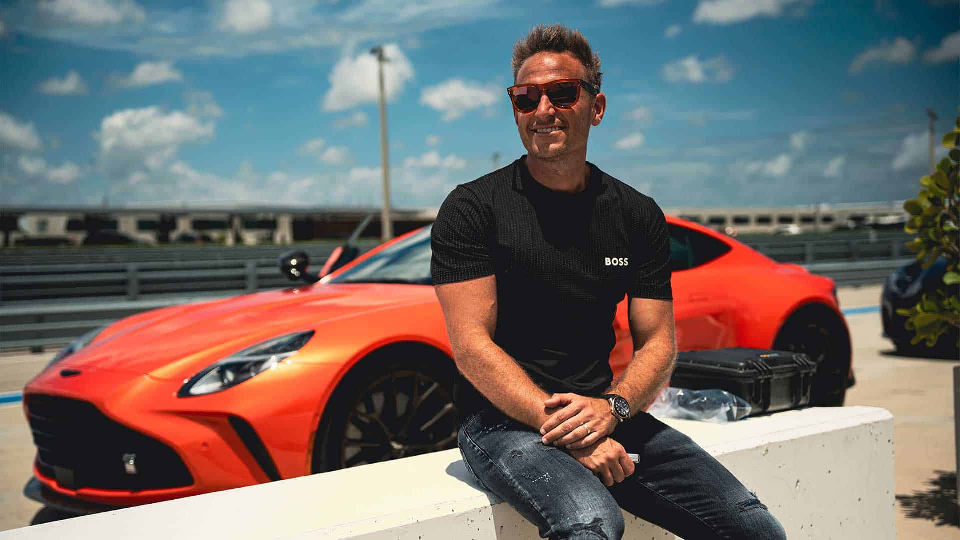 Luxurious red sports car parked outdoors with a smiling man in sunglasses sitting on a white concrete barrier, showcasing high-end automotive lifestyle and business entrepreneurial success.