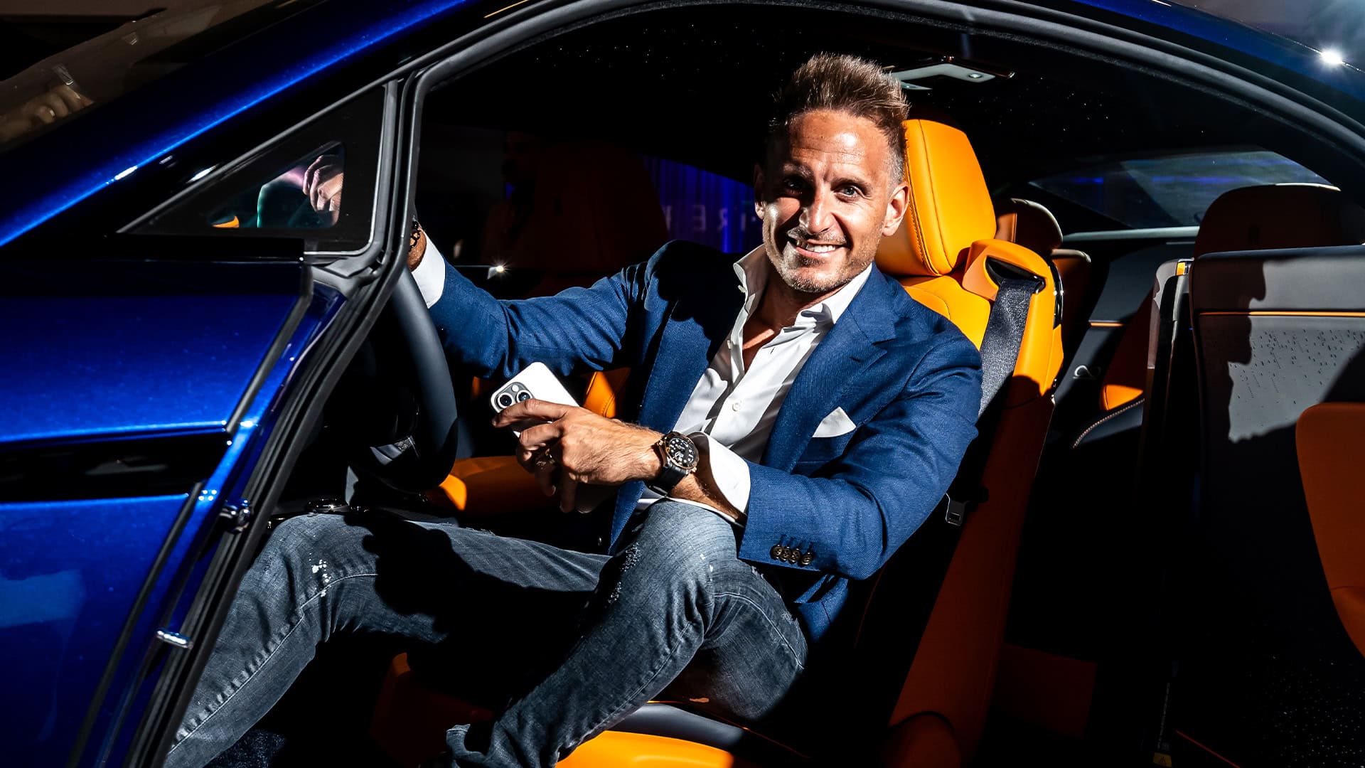 Luxury car interior with a smiling man in a blue suit holding a smartphone, showcasing premium automotive design and lifestyle confidence.
