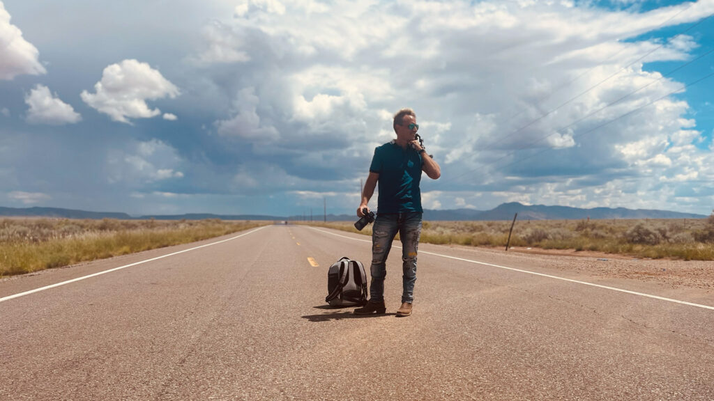 Traveling businessman standing on an open road in desert landscape, carrying a backpack, camera, and phone, under a partly cloudy sky.