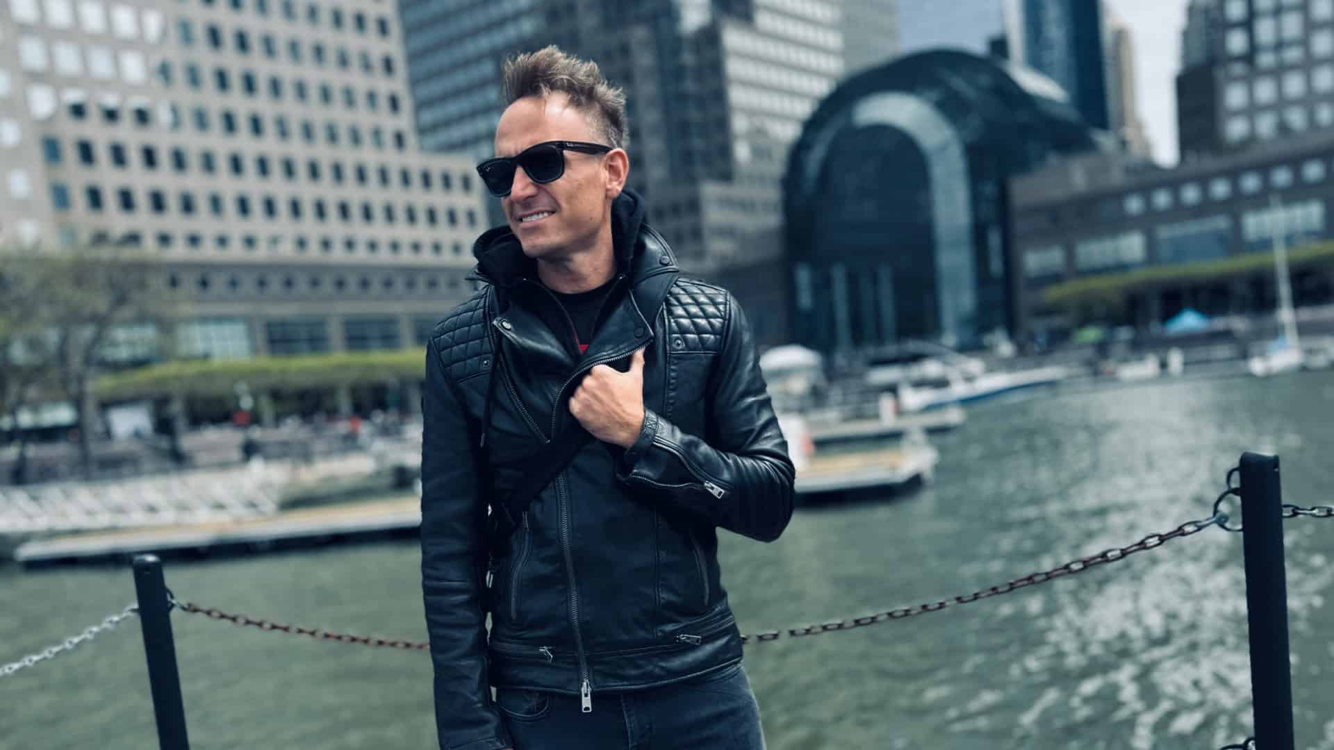 Sleek young man in stylish leather jacket and sunglasses enjoying city waterfront scenery.